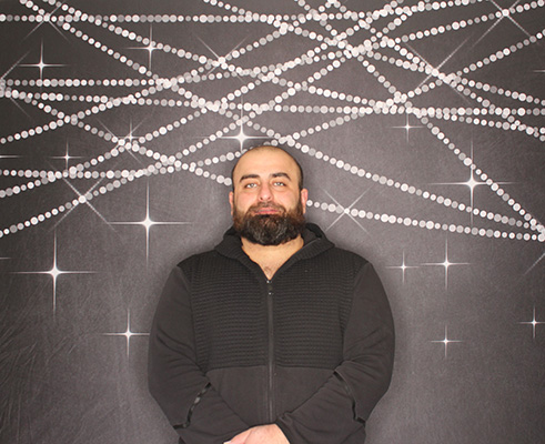 A man with a beard wearing a black zip-up hoodie stands in front of a dark backdrop featuring silver sparkling string lights.